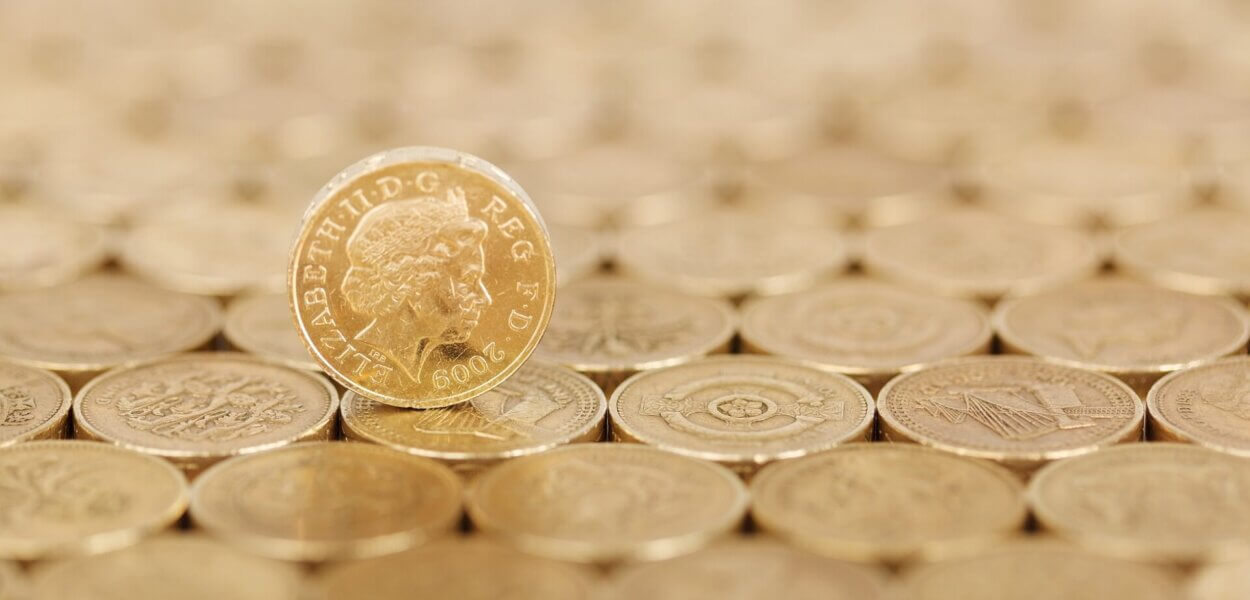 A surface of pound coins with a single coin standing vertically upon it, head side facing towards you