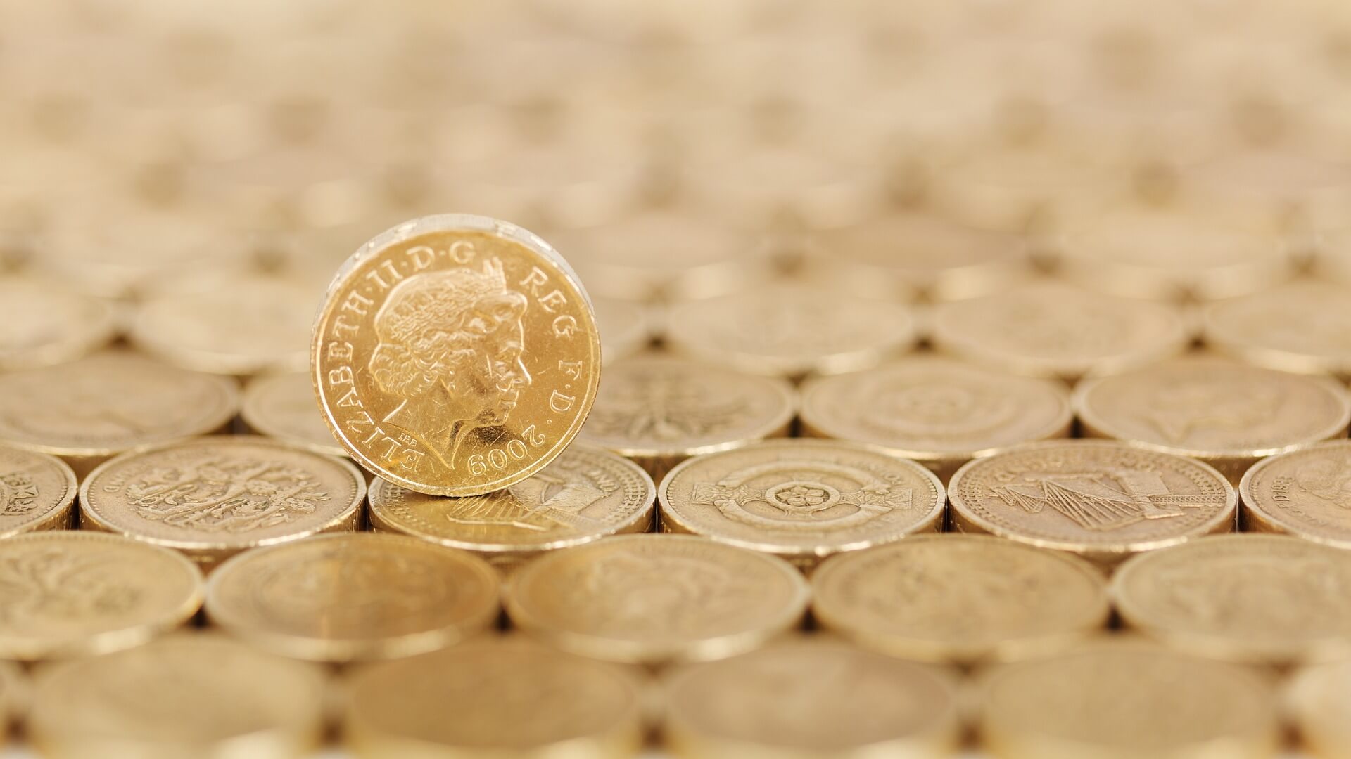 A surface of pound coins with a single coin standing vertically upon it, head side facing towards you